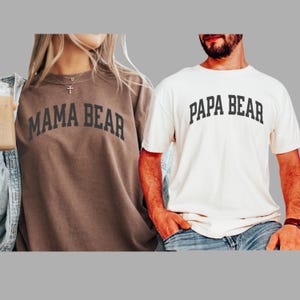May include: Two shirts, one brown and one cream, with the words "MAMA BEAR" and "PAPA BEAR" printed in black, arched across the chest. The brown shirt is a long-sleeved sweatshirt, and the cream shirt is a short-sleeved t-shirt. Both shirts are casual wear.