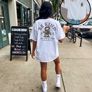 Peut inclure: T-shirt blanc avec un graphique brun d'une personne soulevant un kettlebell avec le texte "BE KIND" et "Get Lots of Steps. Eat Plenty of Protein." La personne porte des baskets blancs et des chaussettes blanches.
