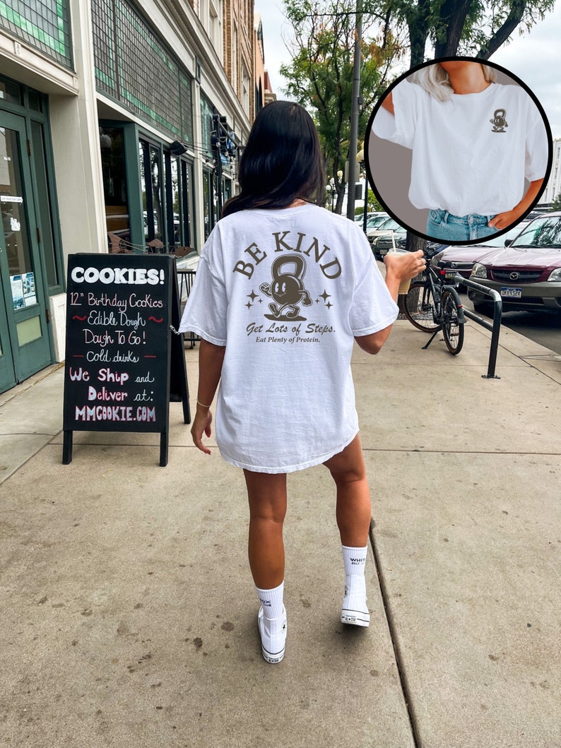 Peut inclure: T-shirt blanc avec un graphique brun d'une personne soulevant un kettlebell avec le texte "BE KIND" et "Get Lots of Steps. Eat Plenty of Protein." La personne porte des baskets blancs et des chaussettes blanches.