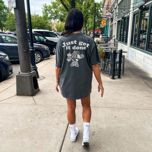 May include: A dark grey t-shirt with the text "Just get it done" and a cartoon character graphic on the back. The person is wearing white socks and trainers. The image is taken outdoors on a pavement.