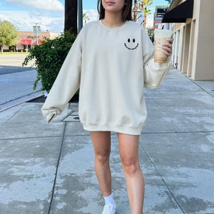 May include: A cream colored sweatshirt with a black smiley face graphic and the text "smile more often" on the front. The person is wearing the sweatshirt and holding a cup of coffee.