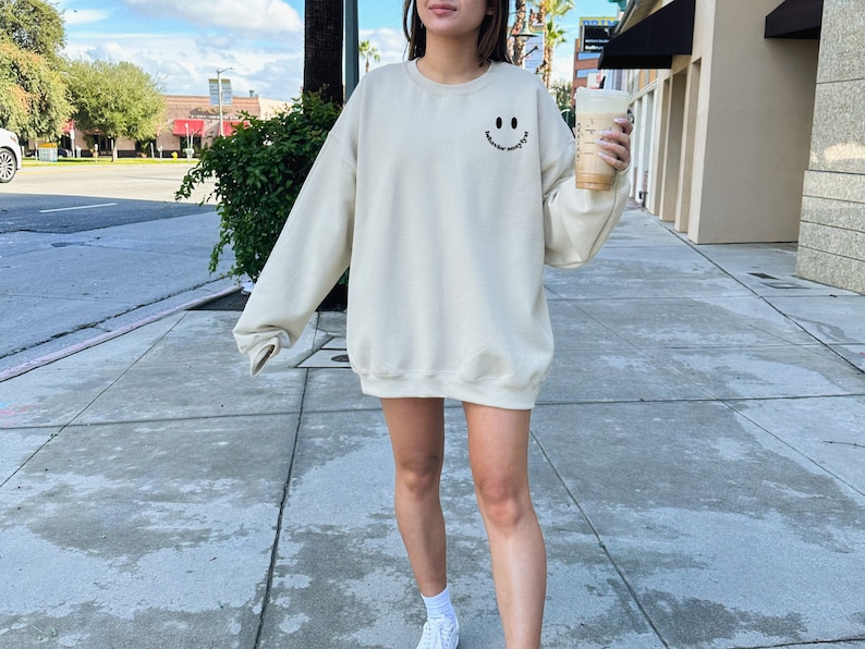 May include: A cream colored sweatshirt with a black smiley face graphic and the text "smile more often" on the front. The person is wearing the sweatshirt and holding a cup of coffee.