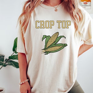 May include: A cream colored crop top with a graphic of corn on the front. The text "CROP TOP" is printed in yellow above the corn graphic.