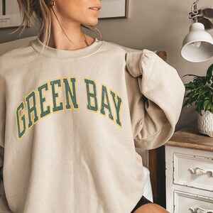 May include: A beige sweatshirt with a green and yellow arched graphic that says "GREEN BAY".