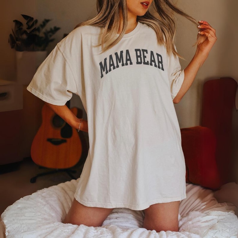 May include: Cream-colored oversized t-shirt with the text "MAMA BEAR" in black, arched across the chest. The shirt has short sleeves and a relaxed fit. The image shows the shirt on a person.