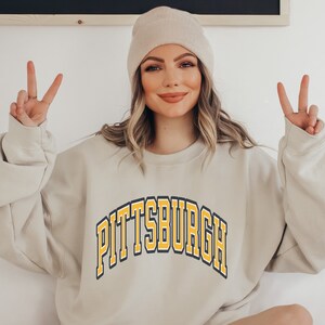 Może przedstawiać: Beżowa bluza z napisem "PITTSBURGH" w ż&oacute;łtych i czarnych łukowatych literach. Bluza jest połączona z beżową czapką. Osoba pokazuje znak pokoju.