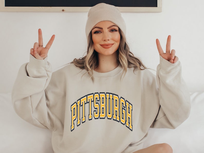 Może przedstawiać: Beżowa bluza z napisem "PITTSBURGH" w ż&oacute;łtych i czarnych łukowatych literach. Bluza jest połączona z beżową czapką. Osoba pokazuje znak pokoju.
