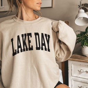 K&ouml;nnte beinhalten: Ein beiger Pullover mit dem Aufdruck "LAKE DAY" in Schwarz.