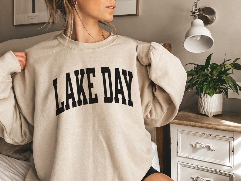 K&ouml;nnte beinhalten: Ein beiger Pullover mit dem Aufdruck "LAKE DAY" in Schwarz.
