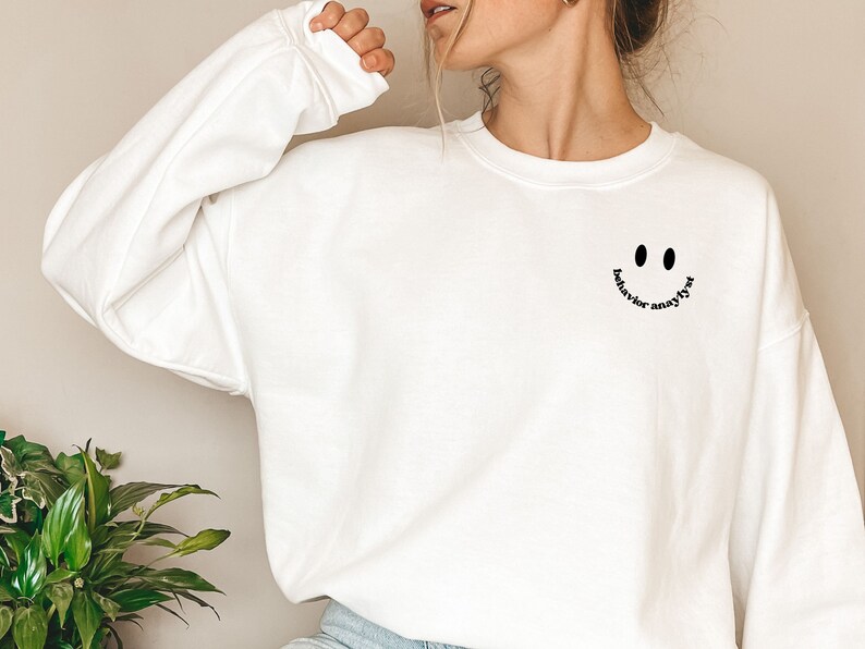 May include: White sweatshirt with a black smiley face and the text "behavior analyst" written around the smile.