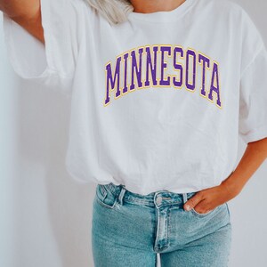 Peut inclure: Un t-shirt blanc avec un graphisme "MINNESOTA" en violet et jaune.