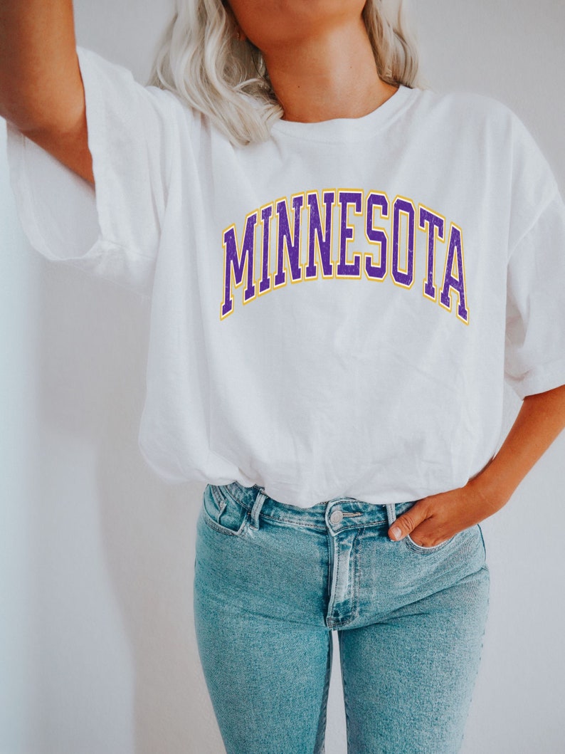 Peut inclure: Un t-shirt blanc avec un graphisme "MINNESOTA" en violet et jaune.