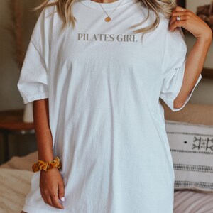 K&ouml;nnte beinhalten: Wei&szlig;es Oversize-T-Shirt mit dem Aufdruck "PILATES GIRL" auf der Vorderseite.
