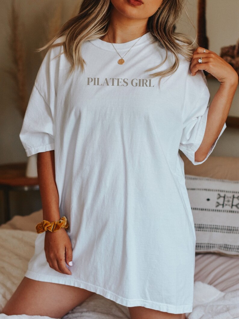 K&ouml;nnte beinhalten: Wei&szlig;es Oversize-T-Shirt mit dem Aufdruck "PILATES GIRL" auf der Vorderseite.