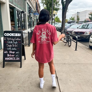 Peut inclure: Une femme portant un t-shirt rouge avec le texte "BE KIND" et un graphique d'une personne tenant un kettlebell. Le texte "Get Lots of Steps" et "Eat Plenty of Potatoes" est &eacute;galement sur le t-shirt.
