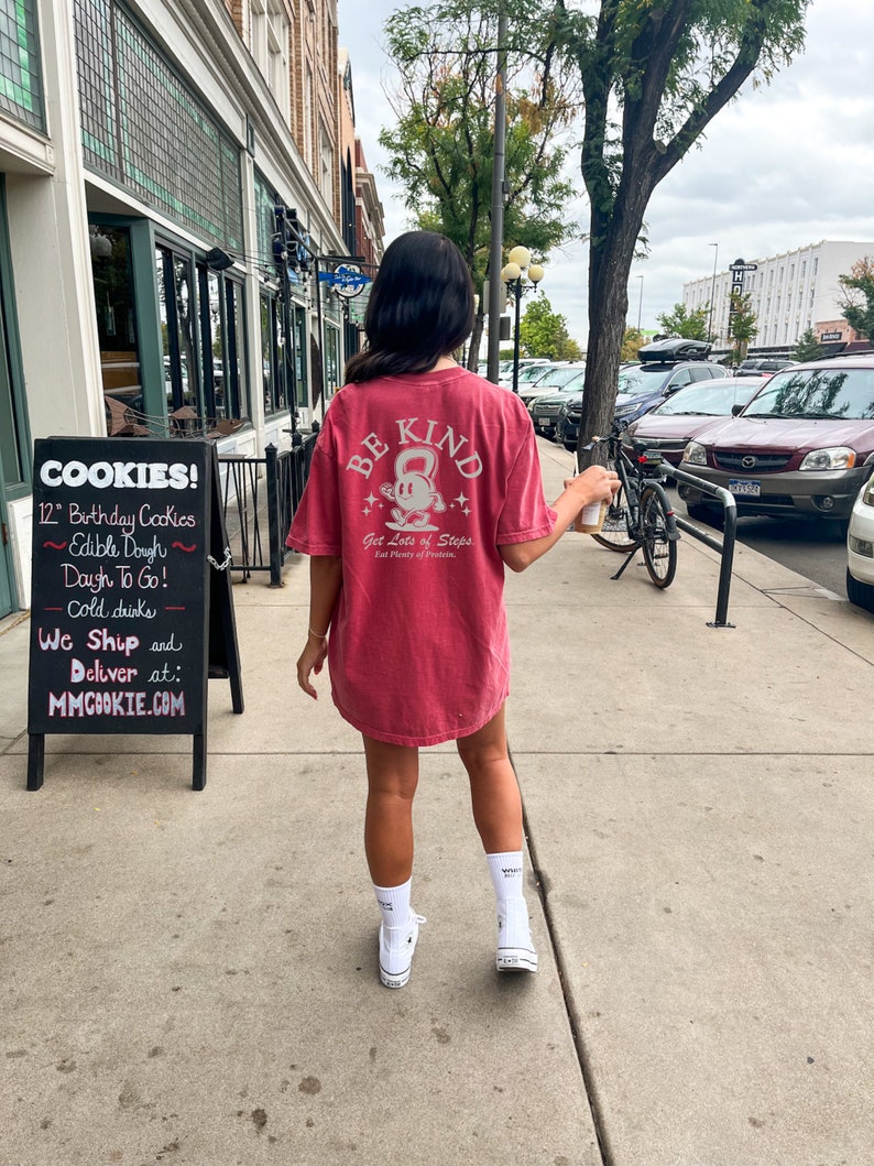 Peut inclure: Une femme portant un t-shirt rouge avec le texte "BE KIND" et un graphique d'une personne tenant un kettlebell. Le texte "Get Lots of Steps" et "Eat Plenty of Potatoes" est &eacute;galement sur le t-shirt.