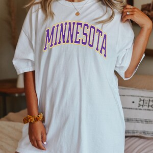 Peut inclure: T-shirt blanc surdimensionn&eacute; avec un graphisme "MINNESOTA" violet et jaune.