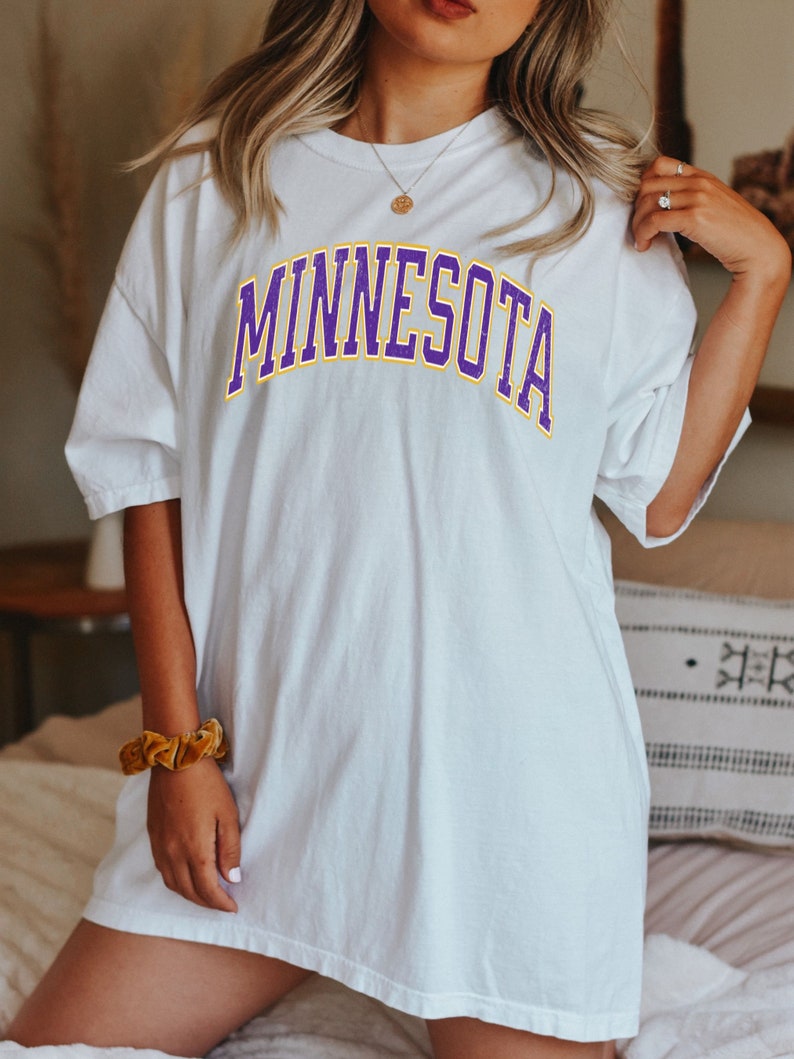 Peut inclure: T-shirt blanc surdimensionn&eacute; avec un graphisme "MINNESOTA" violet et jaune.