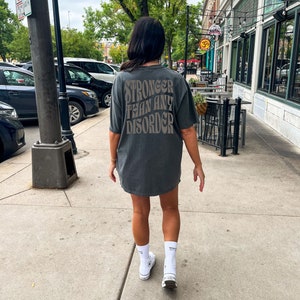 May include: A person wearing a gray t-shirt with the text "Stronger than any disorder" printed on the back. The person is wearing white socks and white sneakers.
