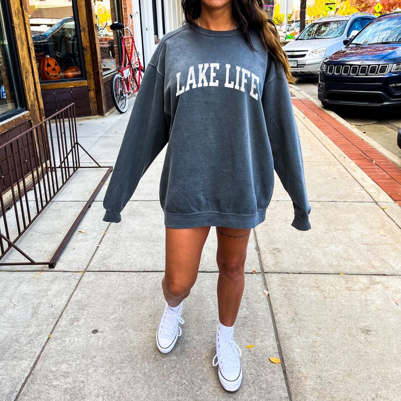 May include: A dark gray sweatshirt with the text "LAKE LIFE" in white lettering. The sweatshirt is oversized and has a relaxed fit. The model is wearing white high-top sneakers.