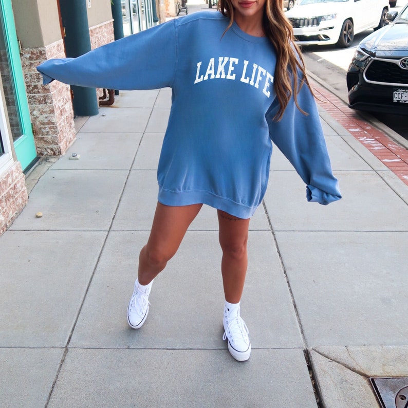 May include: A blue sweatshirt with the text "LAKE LIFE" in white lettering. The sweatshirt is oversized and has a relaxed fit. The model is wearing white high-top sneakers.