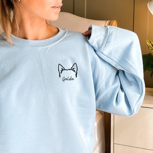 K&ouml;nnte beinhalten: Ein hellblauer Pullover mit einer schwarzen Umrandung eines Hundekopfes und dem Namen "Goldie", der auf der Vorderseite gestickt ist.
