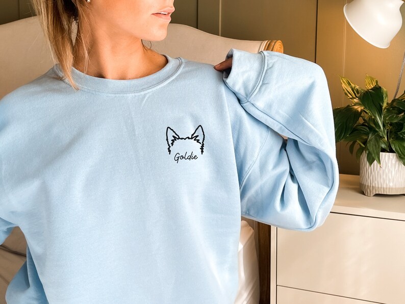 K&ouml;nnte beinhalten: Ein hellblauer Pullover mit einer schwarzen Umrandung eines Hundekopfes und dem Namen "Goldie", der auf der Vorderseite gestickt ist.