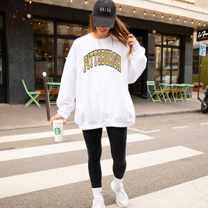 Może przedstawiać: Biała bluza z napisem "PITTSBURGH" w złotych i czarnych literach. Noszona jest czarna czapka z daszkiem z napisem "11:11". Osoba ma na sobie czarne legginsy, białe skarpetki i białe trampki. Trzymany jest kubek Starbucks.