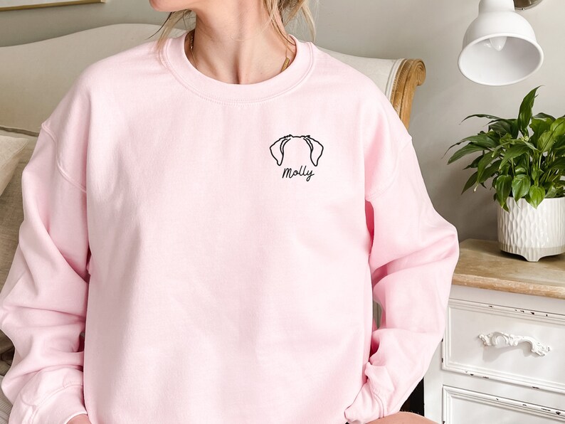 K&ouml;nnte beinhalten: Ein rosa Sweatshirt mit einer schwarzen Linienzeichnung eines Hundekopfes und dem Namen "Molly", der auf der Vorderseite gestickt ist.