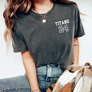 May include: A woman wearing a dark gray t-shirt with the text "TITANS 24" on the front. She is also wearing blue jeans and a gold necklace with a round pendant.