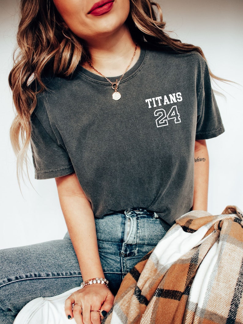 May include: A woman wearing a dark gray t-shirt with the text "TITANS 24" on the front. She is also wearing blue jeans and a gold necklace with a round pendant.
