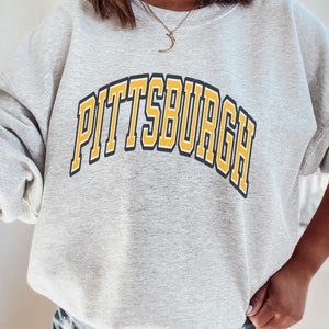 Może przedstawiać: Jasnoszara bluza z napisem "PITTSBURGH" w ż&oacute;łtych literach z czarnym obrysem na piersi. Bluza ma długie rękawy i okrągły dekolt.