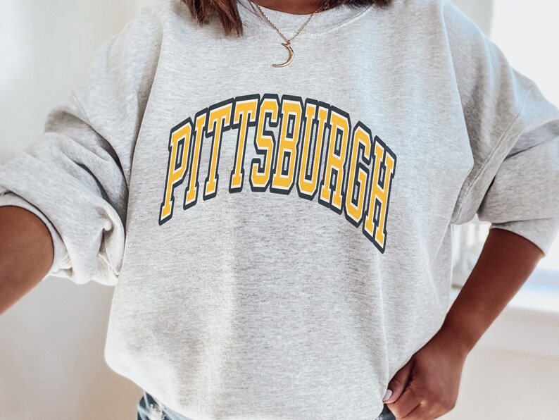 Może przedstawiać: Jasnoszara bluza z napisem "PITTSBURGH" w ż&oacute;łtych literach z czarnym obrysem na piersi. Bluza ma długie rękawy i okrągły dekolt.