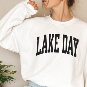K&ouml;nnte beinhalten: Wei&szlig;er Pullover mit schwarzem Text, der "LAKE DAY" in einer College-Schriftart lautet.