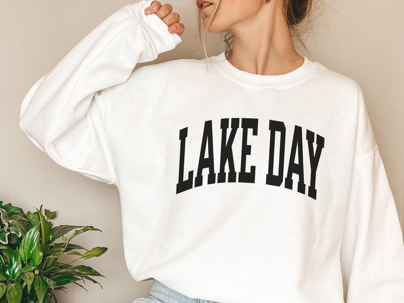 K&ouml;nnte beinhalten: Wei&szlig;er Pullover mit schwarzem Text, der "LAKE DAY" in einer College-Schriftart lautet.