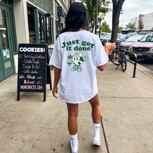 May include: White t-shirt with the slogan "Just get it done" in green. The back of the shirt features a cartoon character holding weights. The person is wearing white socks and trainers. A sign for cookies is visible in the background.