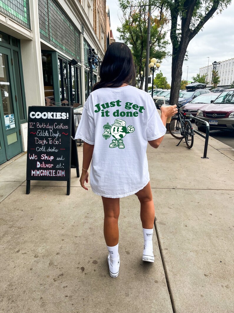 May include: White t-shirt with the slogan "Just get it done" in green. The back of the shirt features a cartoon character holding weights. The person is wearing white socks and trainers. A sign for cookies is visible in the background.