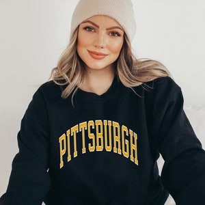 Może przedstawiać: Czarna bluza z napisem "PITTSBURGH" w złotych literach z białym obrysem. Bluza jest noszona z beżową czapką. Osoba się uśmiecha.