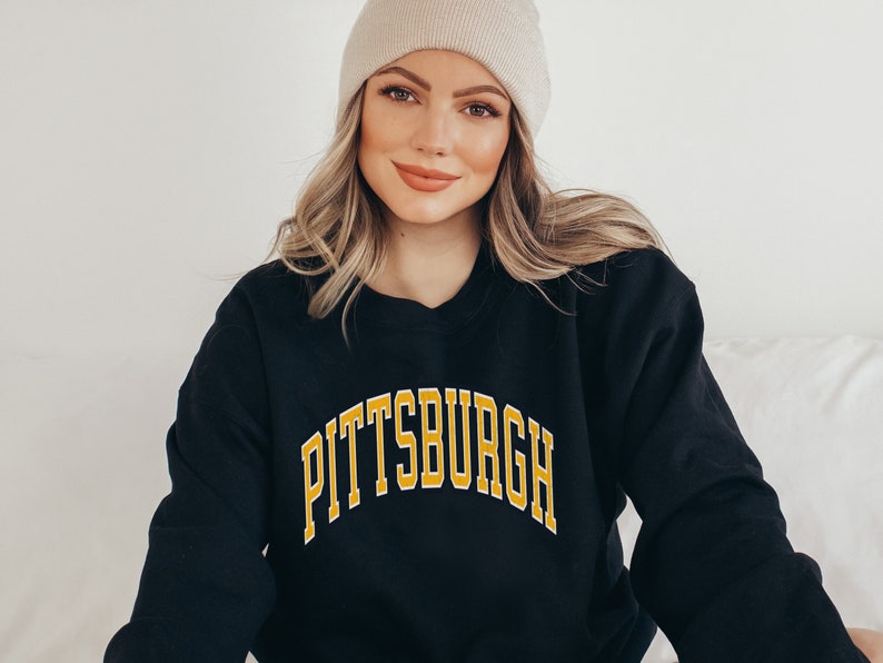 Może przedstawiać: Czarna bluza z napisem "PITTSBURGH" w złotych literach z białym obrysem. Bluza jest noszona z beżową czapką. Osoba się uśmiecha.