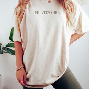 K&ouml;nnte beinhalten: Eine Frau tr&auml;gt ein cremefarbenes, &uuml;bergro&szlig;es T-Shirt mit dem Aufdruck "PILATES GIRL" auf der Vorderseite. Sie tr&auml;gt auch dunkelgr&uuml;ne Radlerhosen.