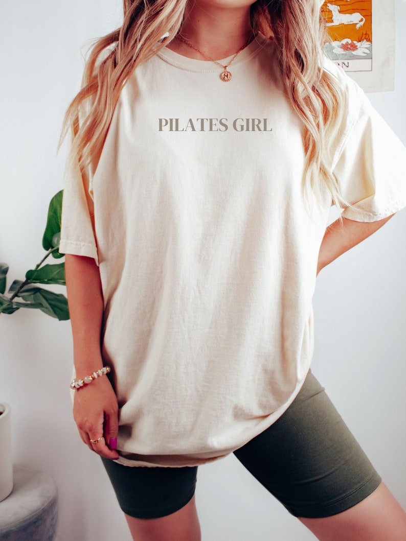 K&ouml;nnte beinhalten: Eine Frau tr&auml;gt ein cremefarbenes, &uuml;bergro&szlig;es T-Shirt mit dem Aufdruck "PILATES GIRL" auf der Vorderseite. Sie tr&auml;gt auch dunkelgr&uuml;ne Radlerhosen.