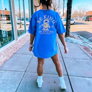 Peut inclure: Un t-shirt bleu avec un motif dor&eacute; d'un personnage de dessin anim&eacute; tenant un kettlebell. Le texte "BE KIND" est au-dessus du personnage et le texte "Get Lots of Steps. Eat Plenty of Protein." est en dessous du personnage.