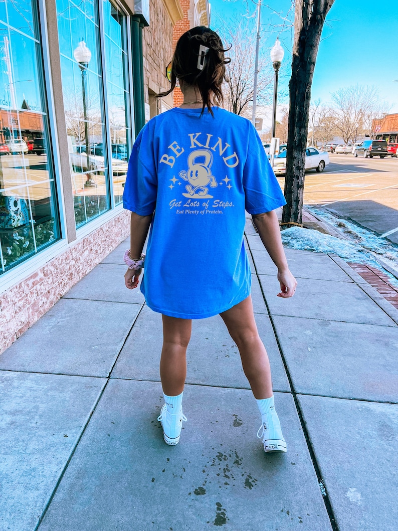 Peut inclure: Un t-shirt bleu avec un motif dor&eacute; d'un personnage de dessin anim&eacute; tenant un kettlebell. Le texte "BE KIND" est au-dessus du personnage et le texte "Get Lots of Steps. Eat Plenty of Protein." est en dessous du personnage.