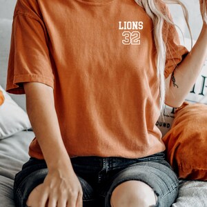 May include: A woman wearing a burnt orange t-shirt with the text "LIONS 32" in white. She is wearing ripped black jeans and sitting on a bed.