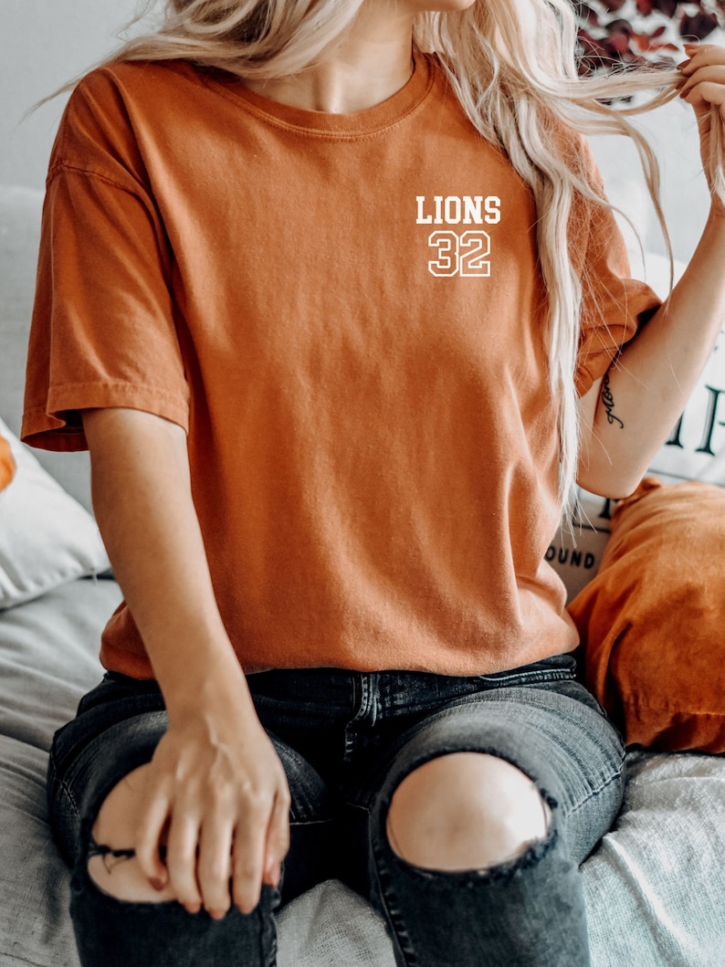 May include: A woman wearing a burnt orange t-shirt with the text "LIONS 32" in white. She is wearing ripped black jeans and sitting on a bed.
