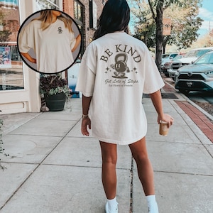 Peut inclure: Un t-shirt blanc avec un graphique brun au dos qui dit "Be Kind" avec un personnage de kettlebell de dessin anim&eacute; et le texte "Get Lots of Steps, Eat Plenty of Protein."
