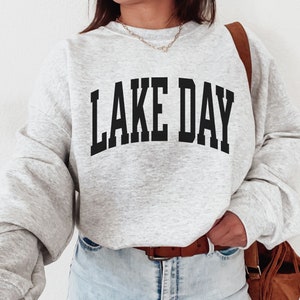 K&ouml;nnte beinhalten: Ein grauer Pullover mit dem Aufdruck "LAKE DAY" in Schwarz auf der Vorderseite.
