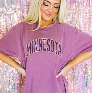 Peut inclure: Un t-shirt surdimensionn&eacute; violet avec le mot "MINNESOTA" imprim&eacute; en lettres jaunes et violettes.