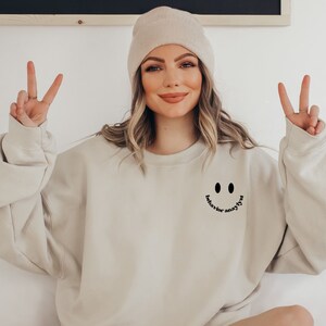 May include: A beige sweatshirt with a black smiley face and the text "behavior for anyone" underneath. The model is wearing a beige beanie and is making a peace sign with both hands.