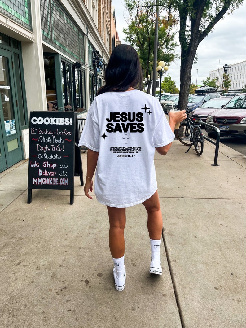 May include: White t-shirt with the text "JESUS SAVES" and a cross printed on the back. The text "FOR GOD SO LOVED THE WORLD THAT HE GAVE HIS ONE AND ONLY SON, THAT WHOSOEVER BELIEVES IN HIM SHALL NOT PERISH BUT HAVE ETERNAL LIFE." and "JOHN 3:16-17" are also printed on the back.